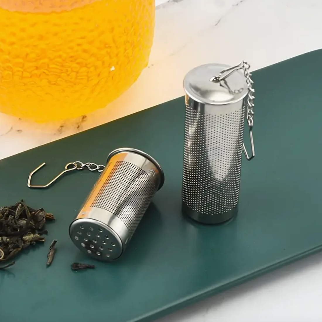 Two metal tea infusers on a green tray with loose tea leaves and a glass of tea in the background.